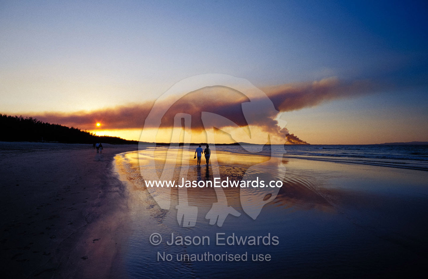 A couple walking on the beach at sunset, with a distant bush fire. Noosa, Queensland, Australia.