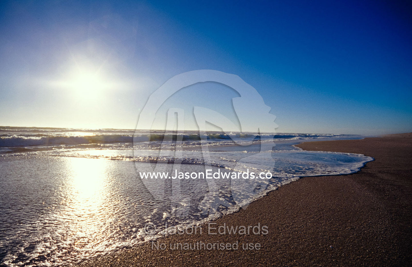 Gentle waves lap onto a pristine sandy beach with the sun reflecting. Coorong National Park, Younghusband Penninsula, South Australia.