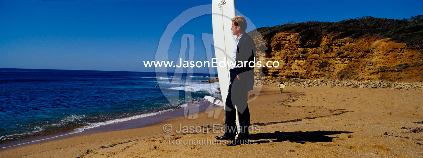 A businessman in a suit waits on the beach to go longboard surfing. Bells Beach, Victoria, Australia.