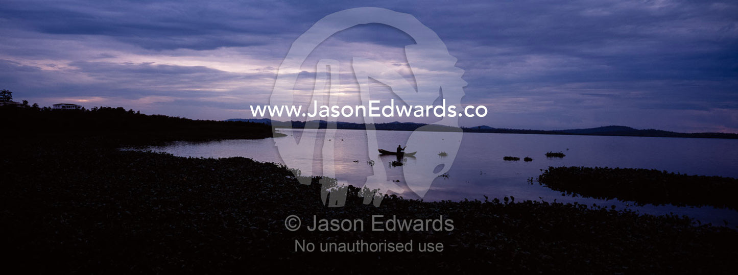 A fisherman in a dugout canoe casts a pole on Lake Victoria at dawn. Lake Victoria, Kampala, Uganda.