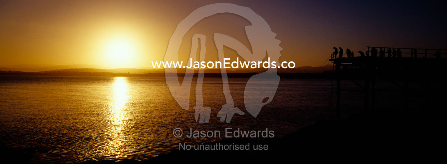 Holiday makers and fishermen in silhouette crowd a jetty at sunset. Merimbula, New South Wales, Australia.