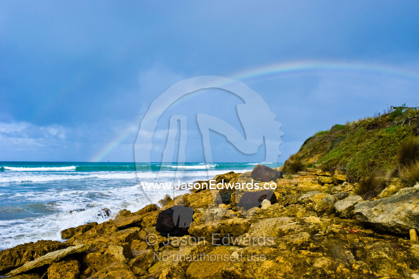 A rainbow rises out of the ocean near a boulder-strewn shoreline. Point Impossible, Victoria, Australia.