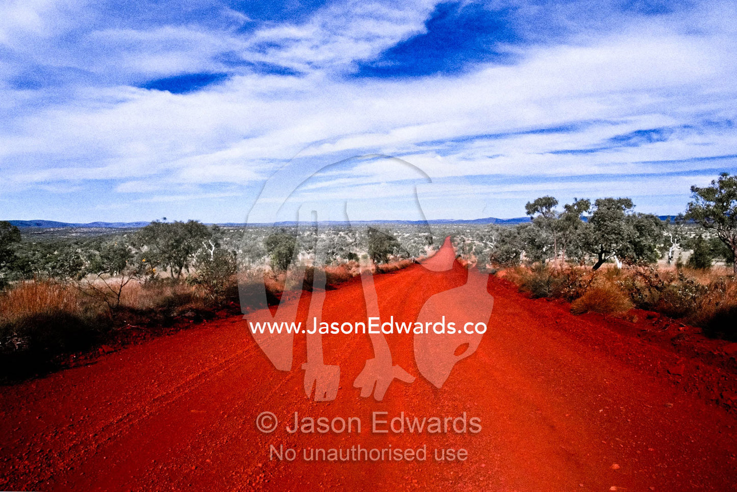 A rich red volcanic track runs straight across the desert plain. Karijini National Park, Western Australia, Australia.