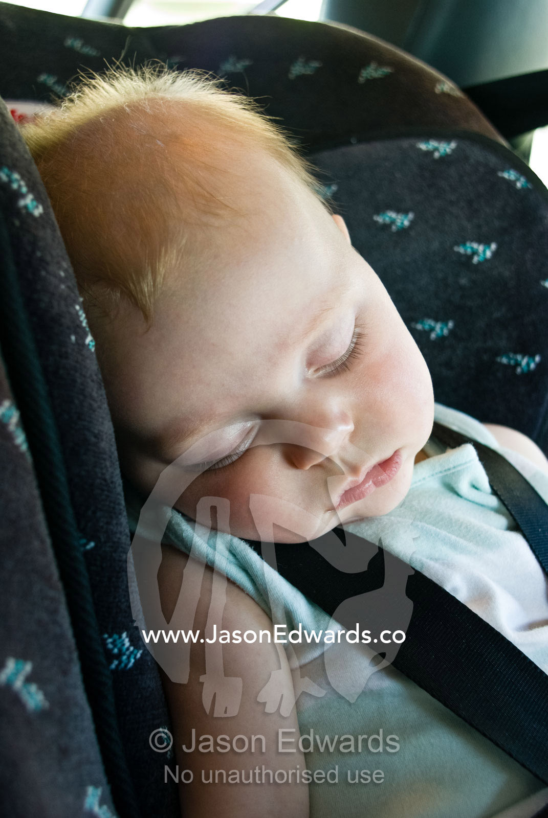 An angelic fair-haired baby exhausted and fast asleep in a car seat. Carlton, Victoria, Australia.