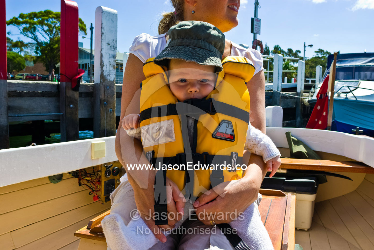 A small baby in an oversized life jacket sails onto a lake in a boat. Metung, Gippsland Lakes, Victoria, Australia.