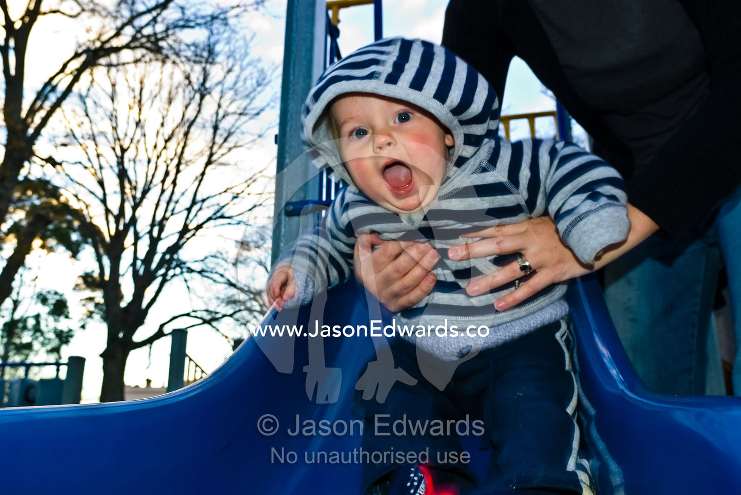 An excited baby shoots safely down a park slide in his mothers hands. Curtain Square, North Carlton, Victoria, Australia.