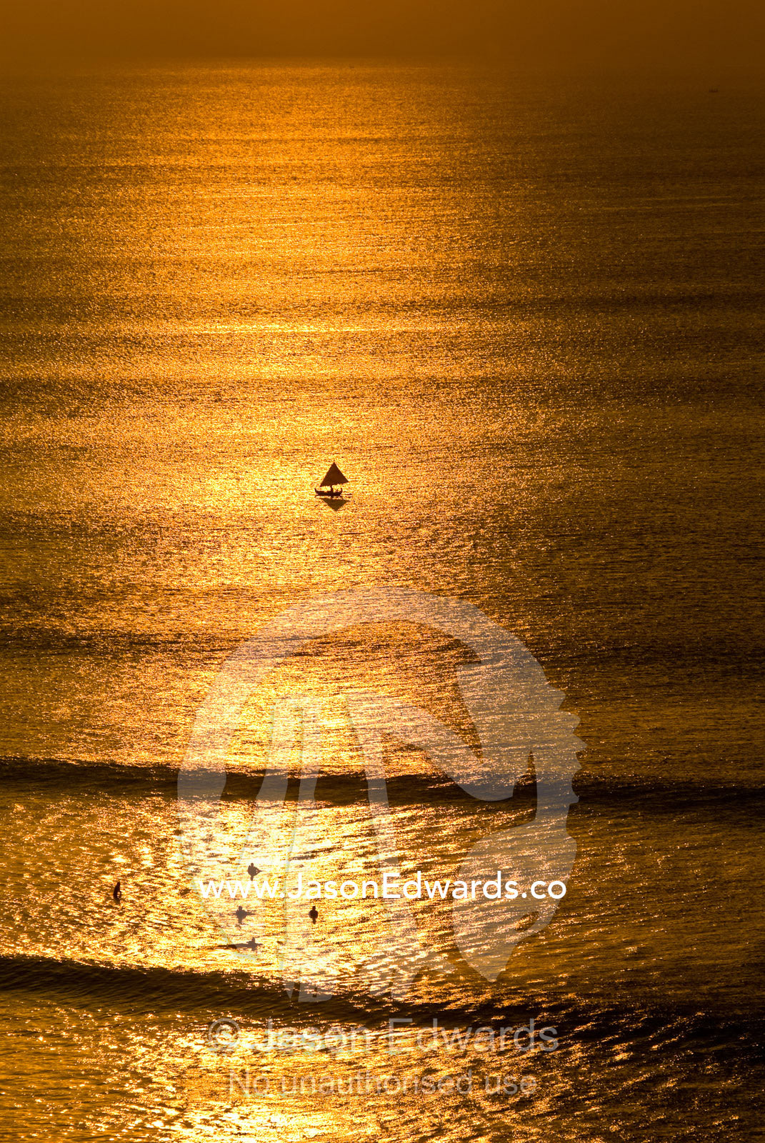 A small fishing boat heads out to sea at sunset past surfers. Indian Ocean, Bukit Peninsula, Bali, Indonesia.