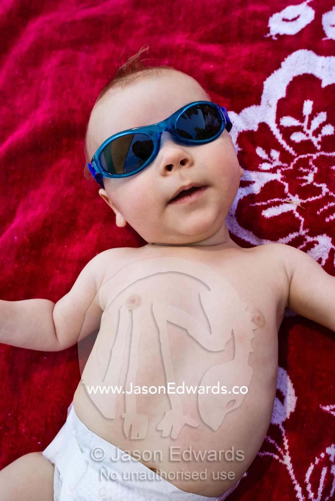 A cool baby in sunglasses relaxes on a beach towel on a summers day. Carlton Baths, Carlton, Victoria, Australia.