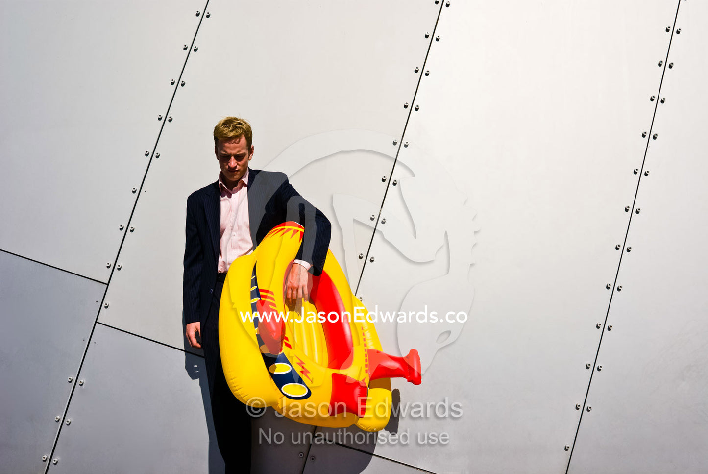 A businessman rests against a stainless steel wall with a toy boat. Carlton Gardens, Victoria, Australia.