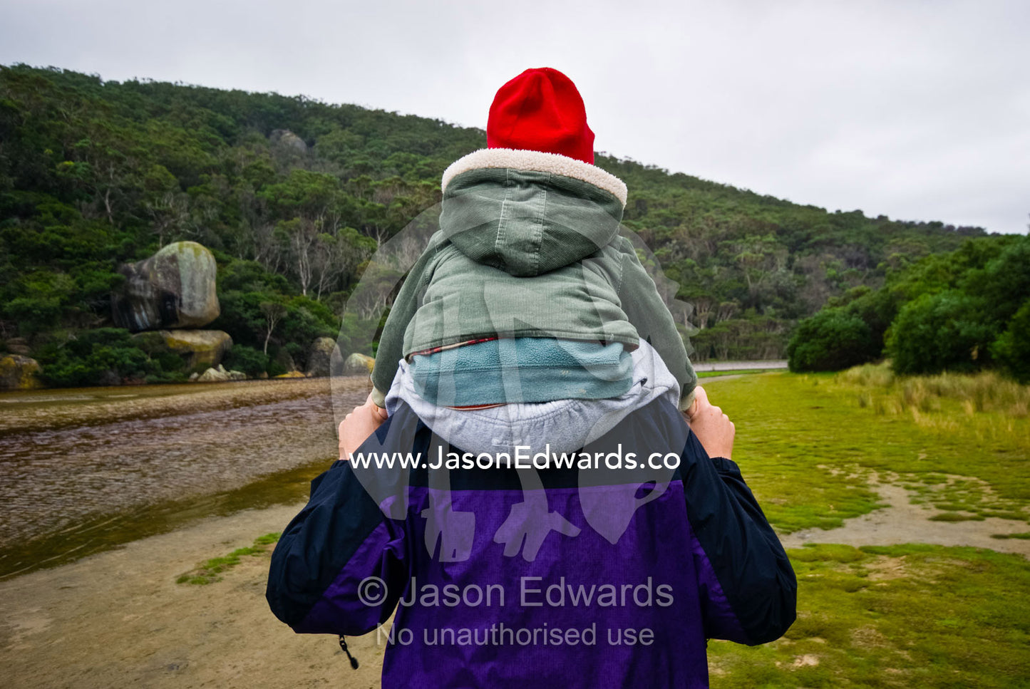 A small boy rides on his mothers shoulders whilst hiking by a river. Tidal River, Wilsons Promontory National Park, Victoria, Australia