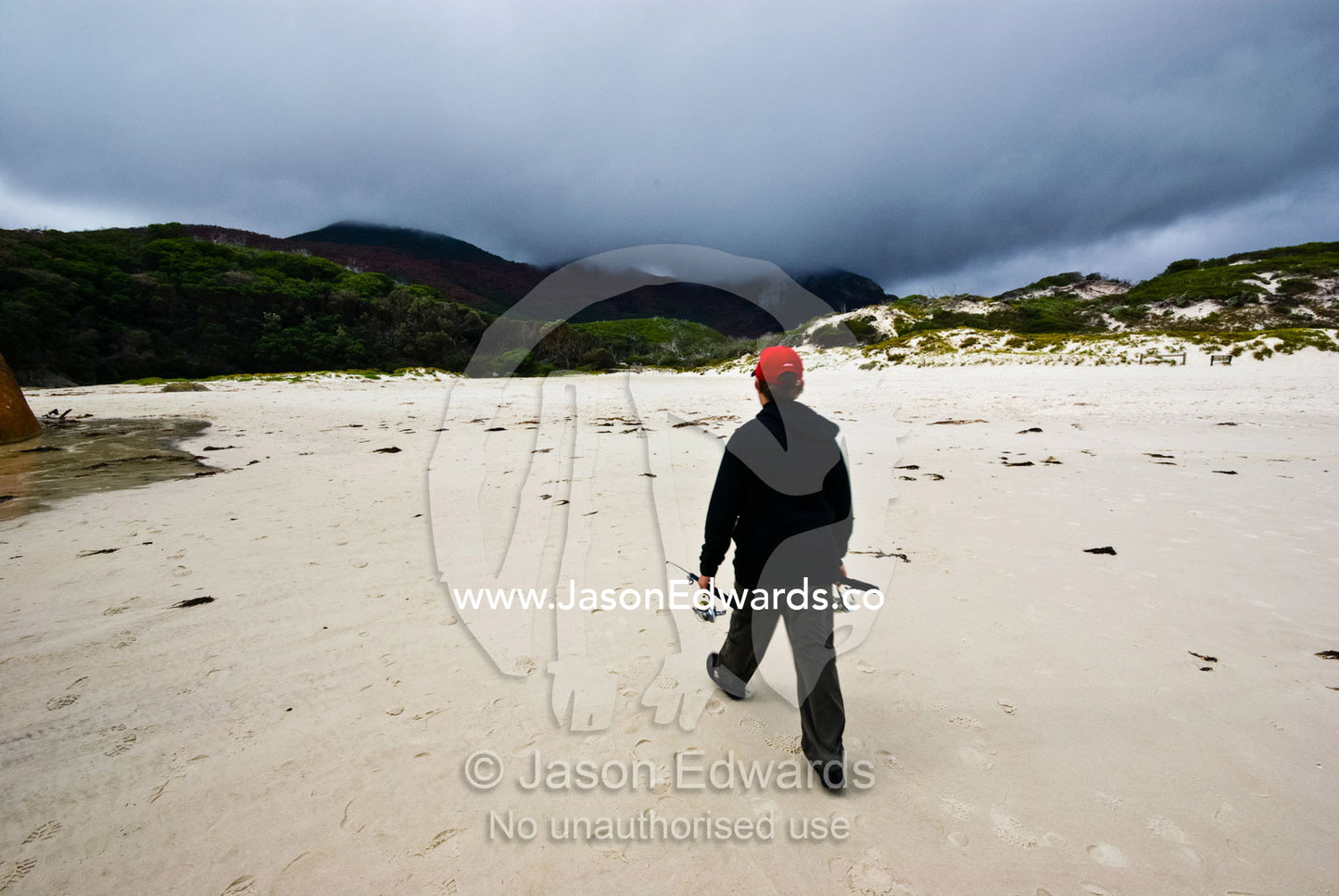 A teenage boy walks along an empty beach with his fishing rods. Squeaky Beach, Wilsons Promontory National Park, Victoria, Australia
