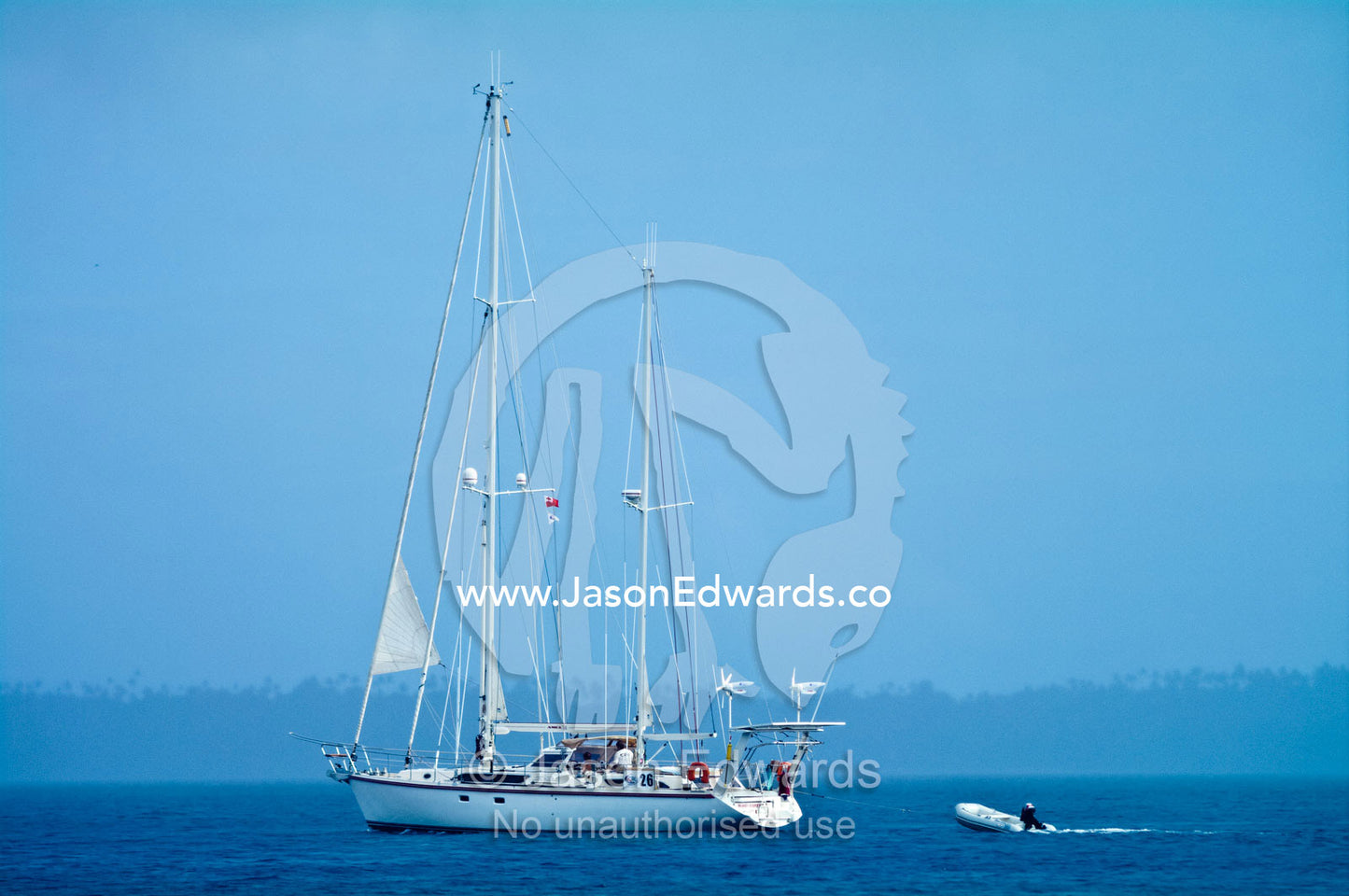 An ocean yacht sails past a lush tropical island under motor power. Vava'u Island Group, Pacific Ocean, Oceania, Kingdom of Tonga.