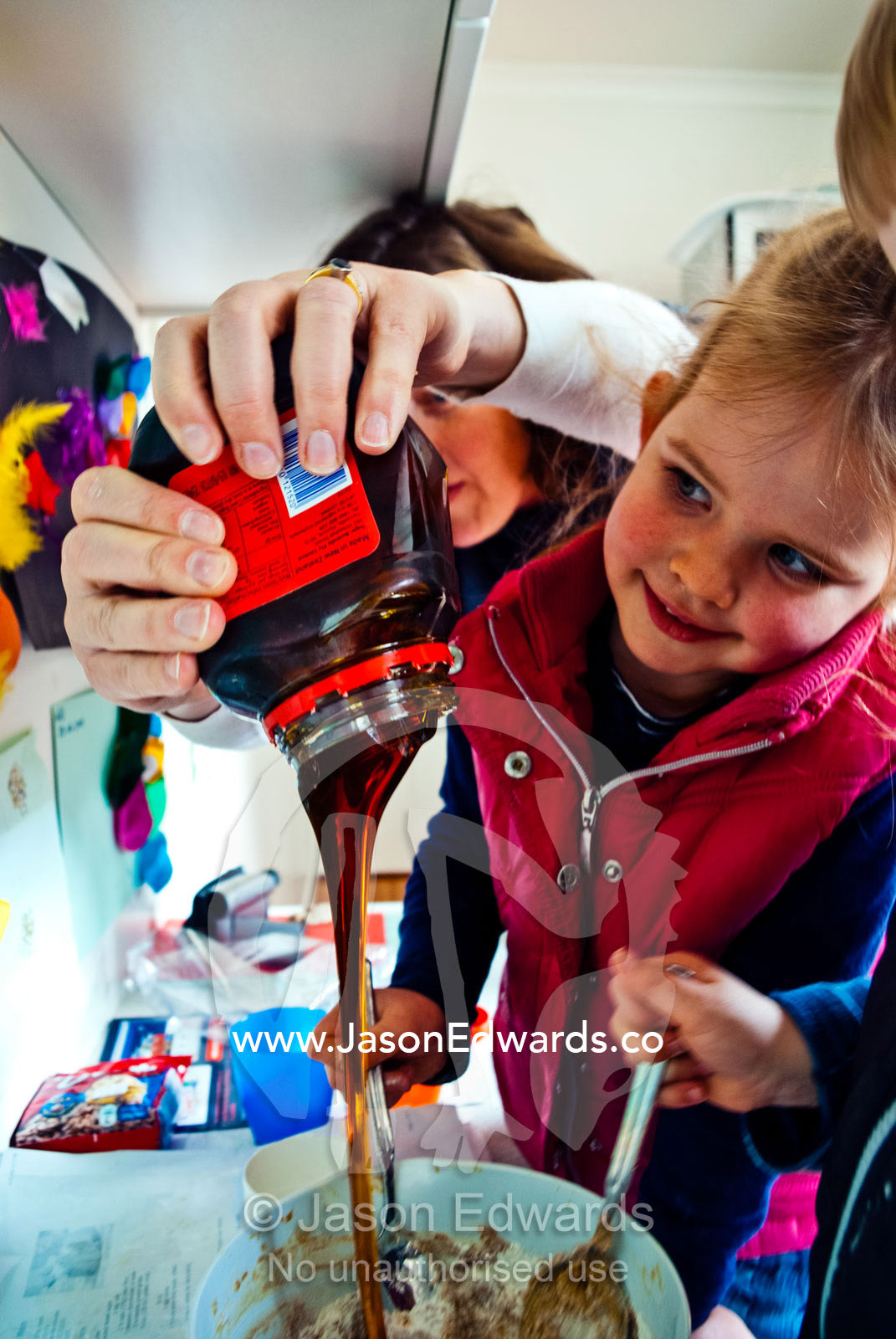 A mother helps children bake gingerbread by pouring the golden syrup. Carlton North, Melbourne, Victoria, Australia.