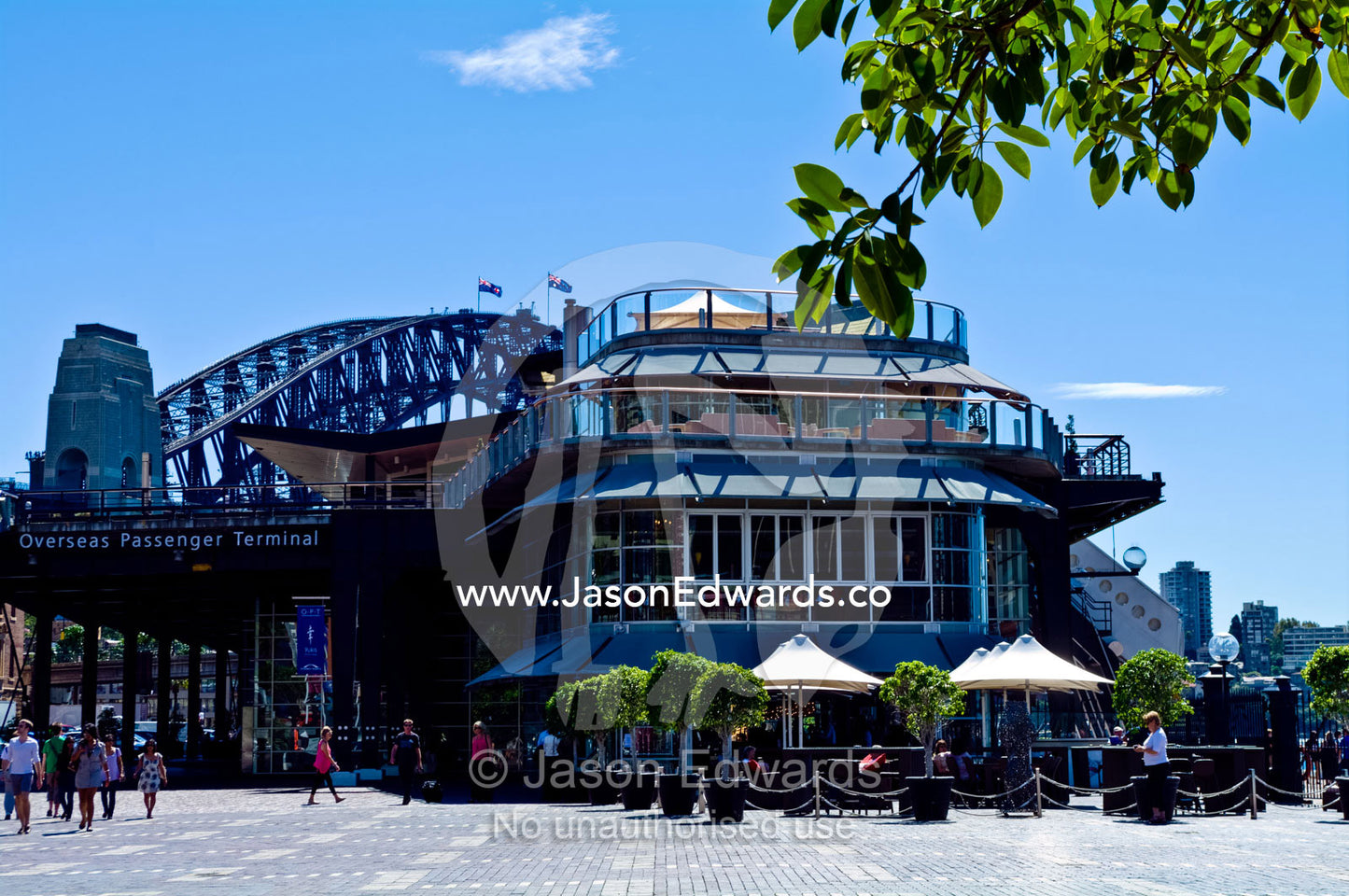 The Sydney Harbour Bridge rises behind restuarants at a ferry port. Sydney Harbour, Sydney Cove, Warrane, Port Jackson, Circular Quay, Sydney, New South Wales, Australia.