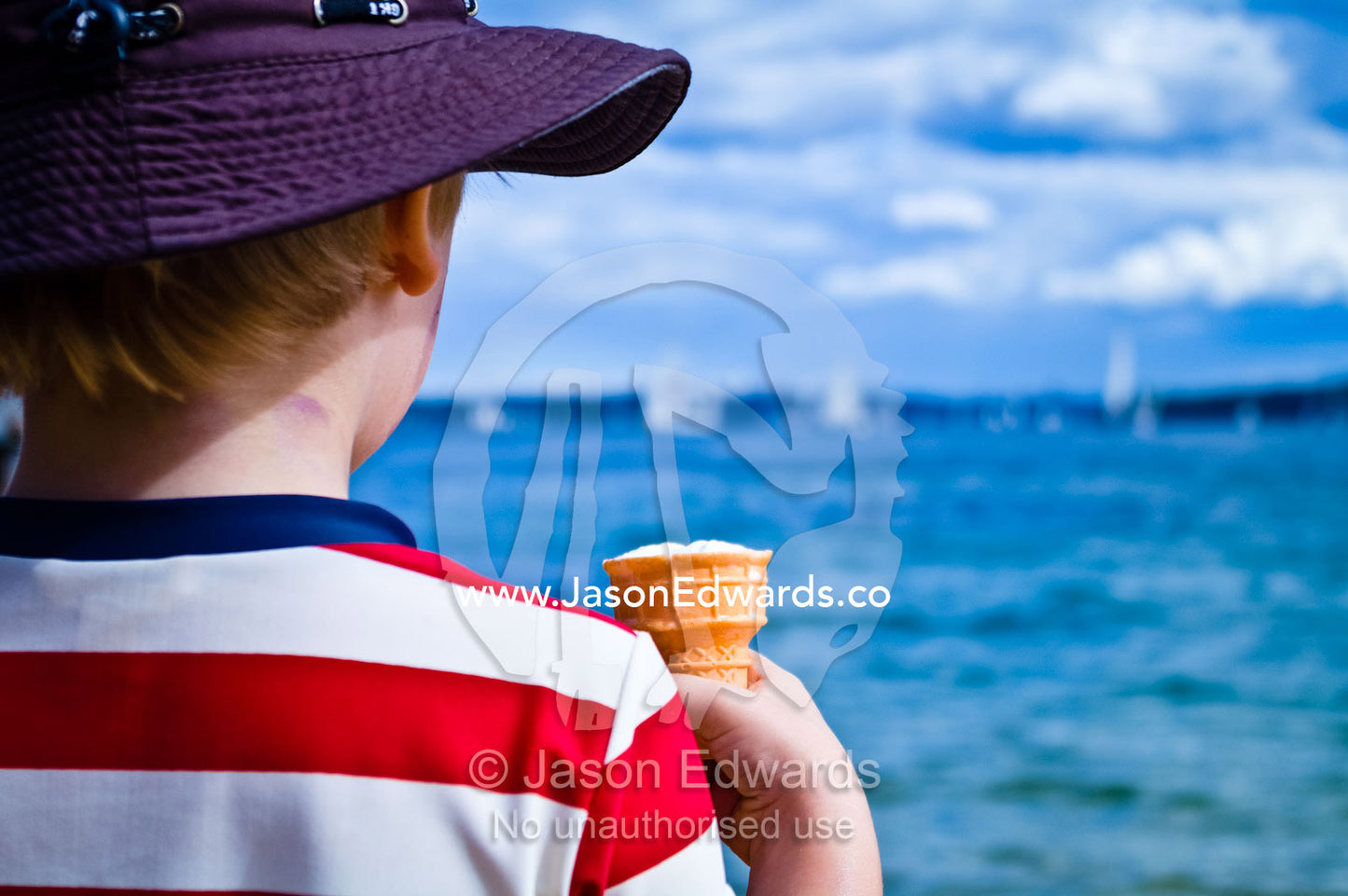 A young boy eats an ice-cream cone whilst watching yachts sailing. Manly Wharf, Manly Cove, Manly, Sydney Harbour, Sydney, New South Wales, Australia.