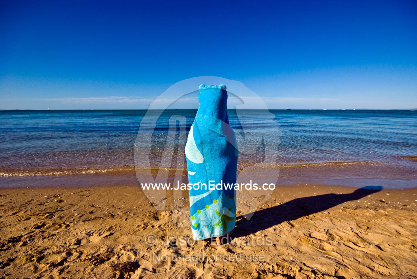 A boy on the beach watching the ocean wrapped in an octopus towel. Rye Beach, Melbourne, Victoria, Australia.
