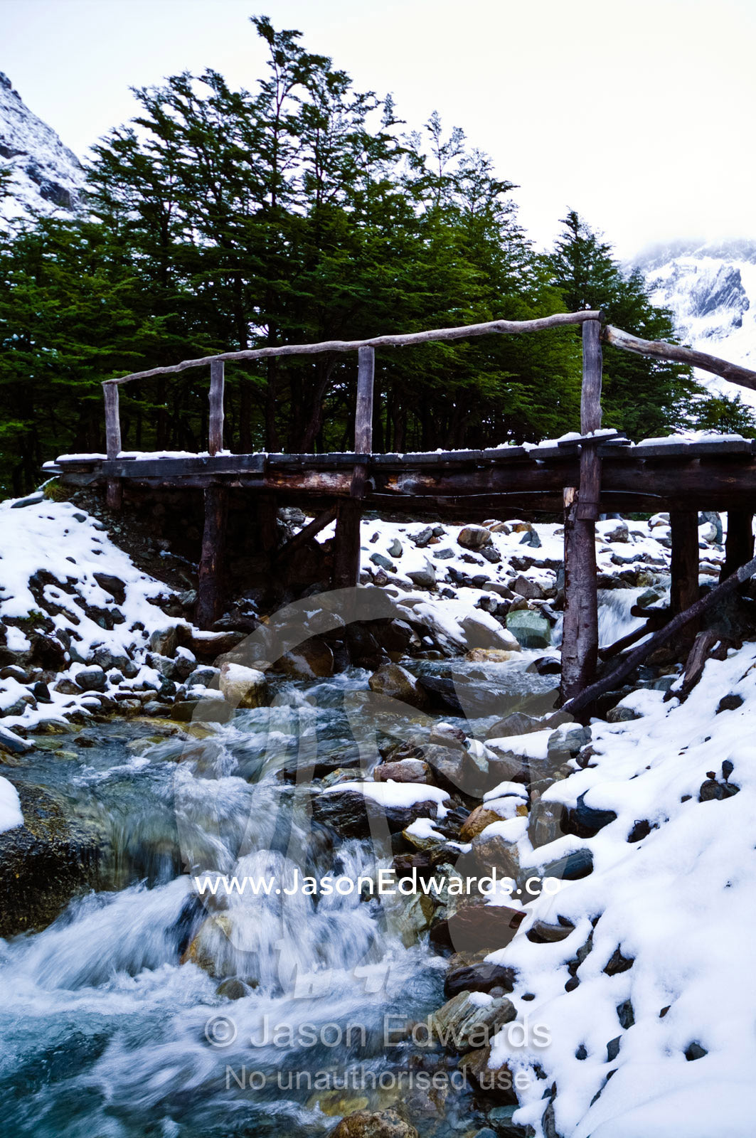 A log and timber footbridge over a stream in a snowy mountain valley. Glacier El Martial, Ushuaia, Fuegian Andes, Tierra del Fuego Archipelago, Straits of Magellan, Tierra del Fuego Province, Patagonia, Argentina.