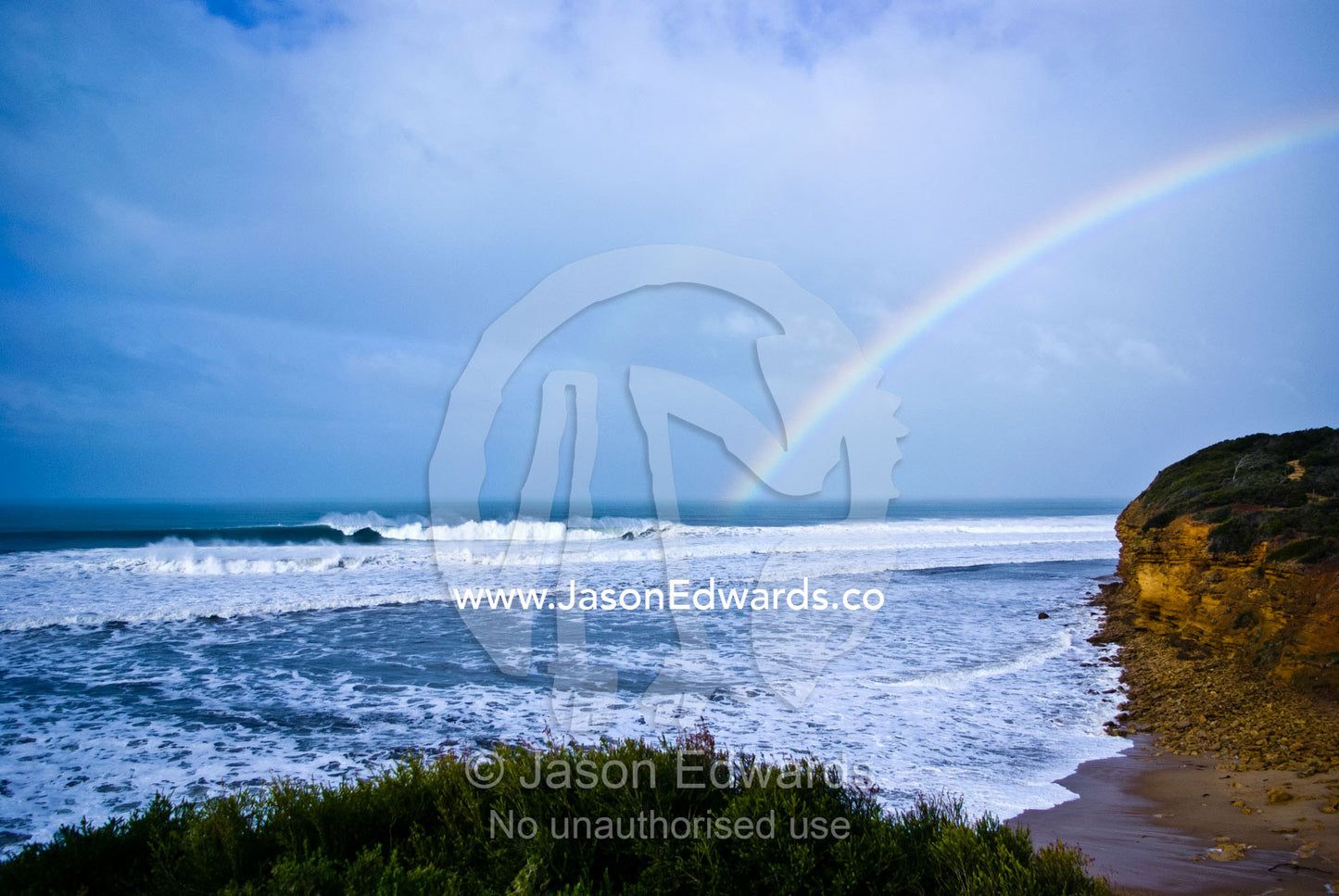 An iridescent rainbow touches the ocean and huge waves at Bells Beach. Bells Beach Surfing Recreation Reserve, Surf Coast, Victoria, Australia.