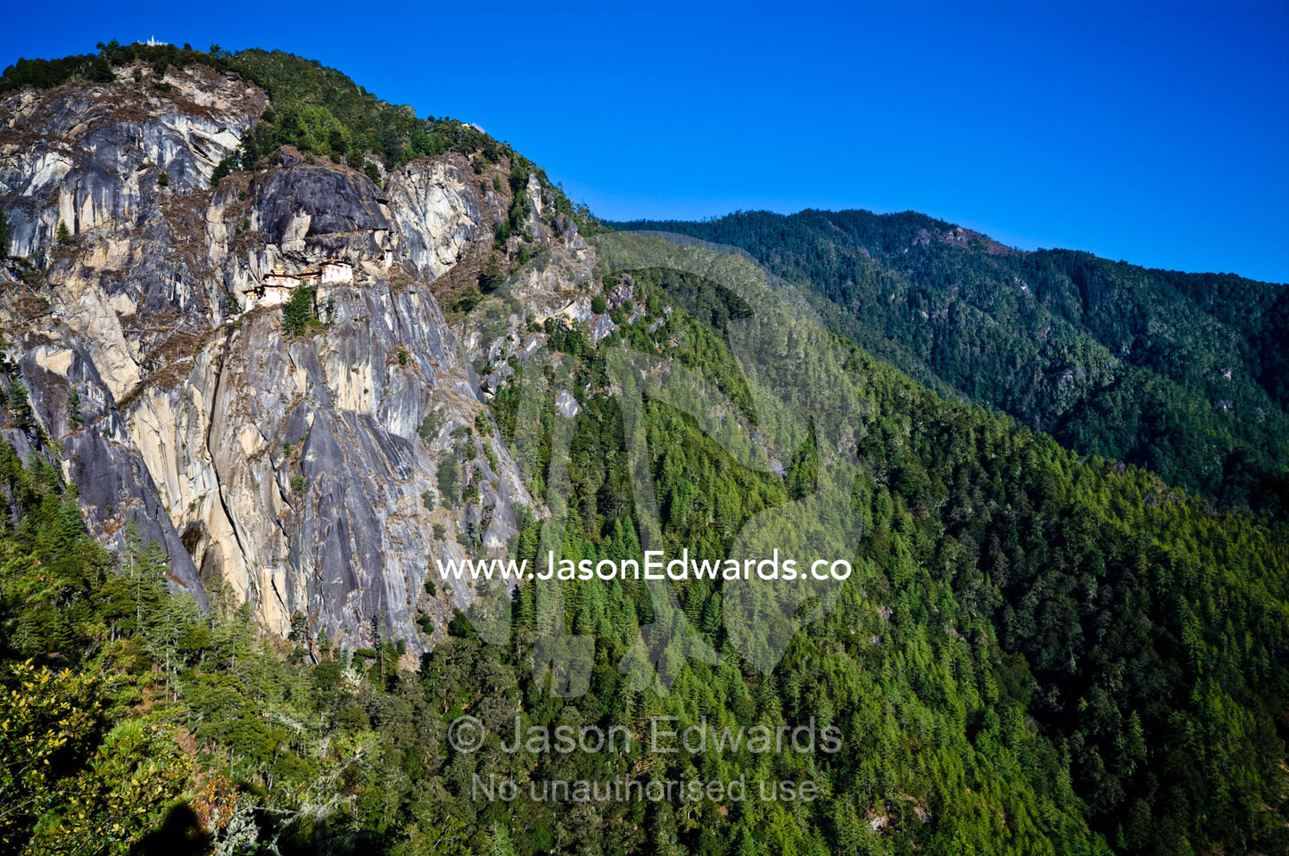 An ancient Buddhist monastery perched on a sheer cliff face above a forested valley in the Himalaya. Paro Taktsang, The Tigers Nest, Paro Valley, Paro District, Bhutan.