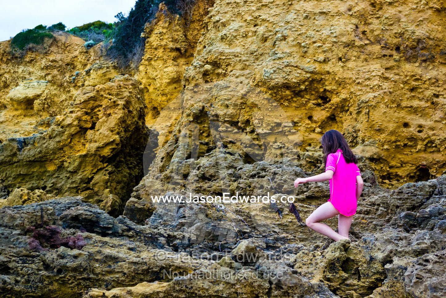 A girl clambers over rugged sharp rocks beneath a cliff at the beach. Torquay, Surf Coast, Victoria, Australia.