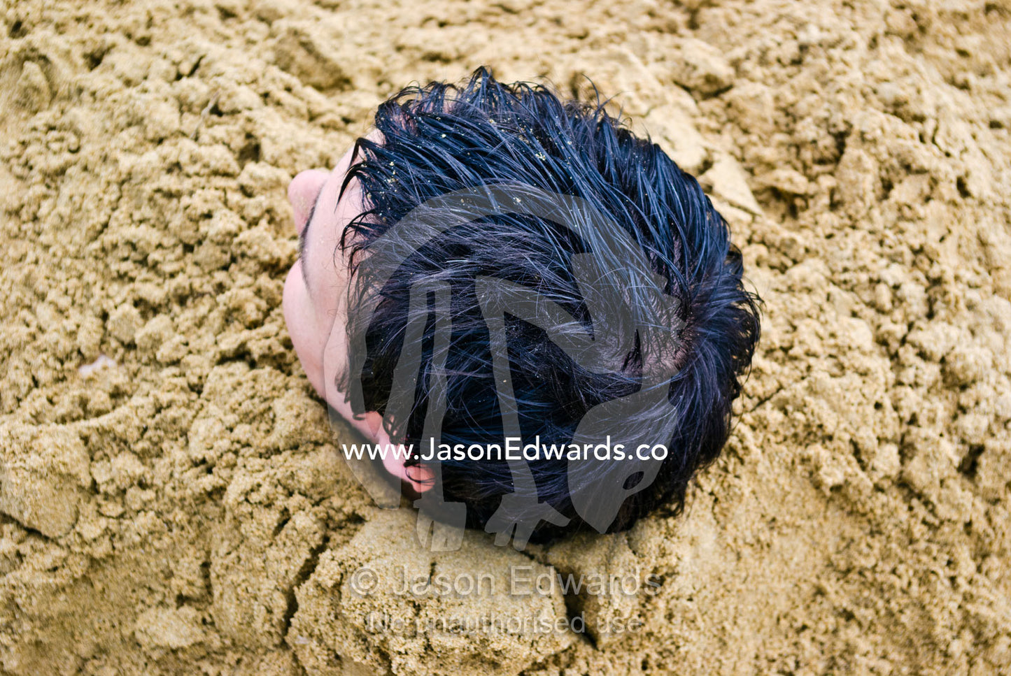 A mans head emerges from the sand buried up to his neck on the beach. Torquay, Surf Coast, Victoria, Australia.