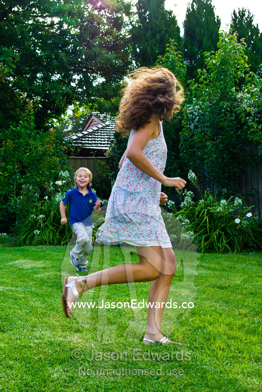 A young boy chases a teenage girl running and playing in a backyard. Malvern East, Melbourne, Victoria, Australia.