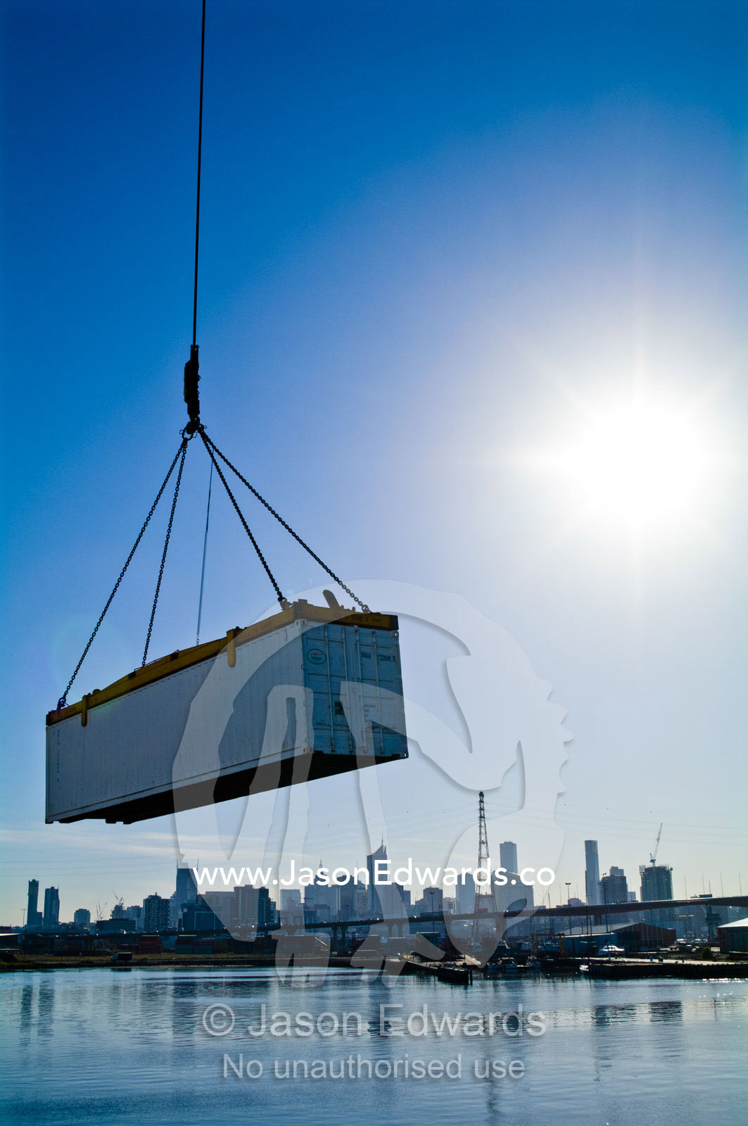 A crane lifts a shipping container over a river past a city skyline. Yarra River, Appleton Dock, Port of Melbourne, West Melbourne, Victoria, Australia.