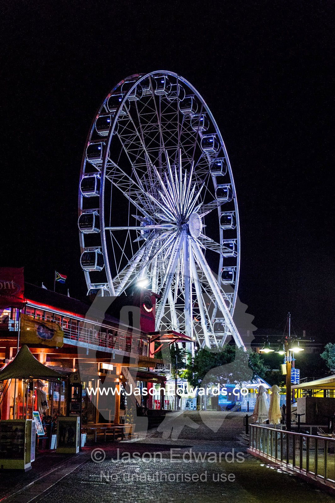 Ferris wheel illuminated by neon lights in a marina shopping district. Cape Town, Victoria and Alfred Waterfront, Western Cape, South Africa.
