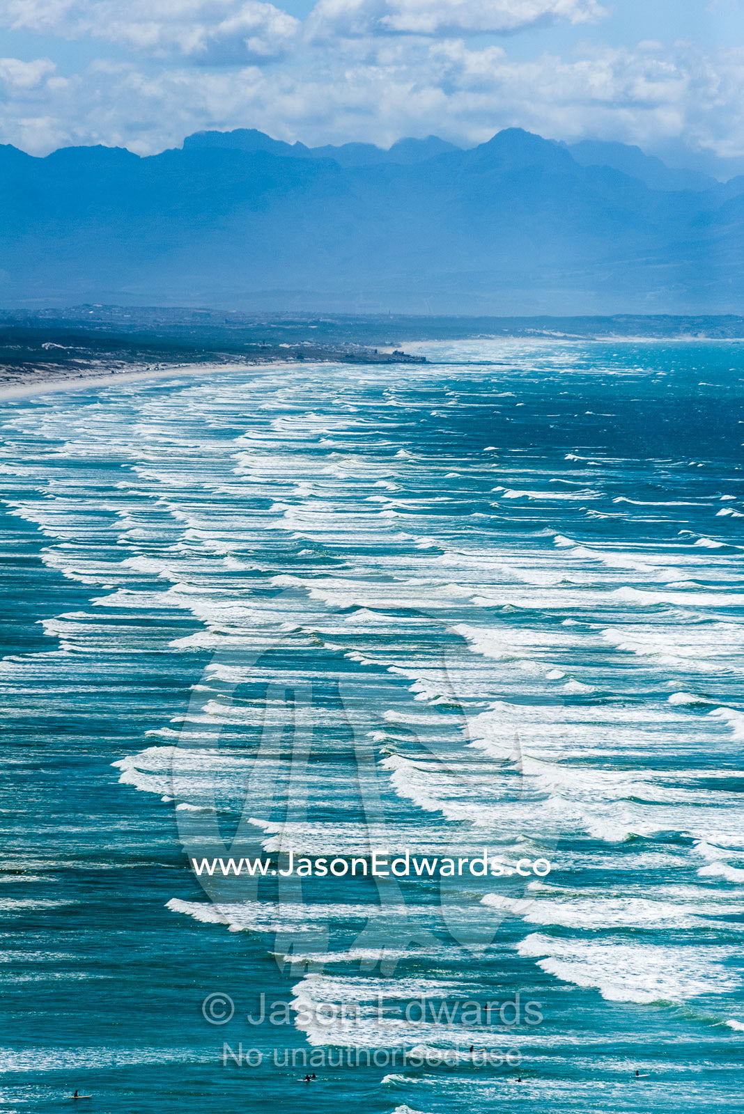 Endless waves roll onto a long, white sandy beach under a summer sky. Surfers Corner, Muizenberg, Cape Town, Chapmans Peak Drive, Western Cape, South Africa.