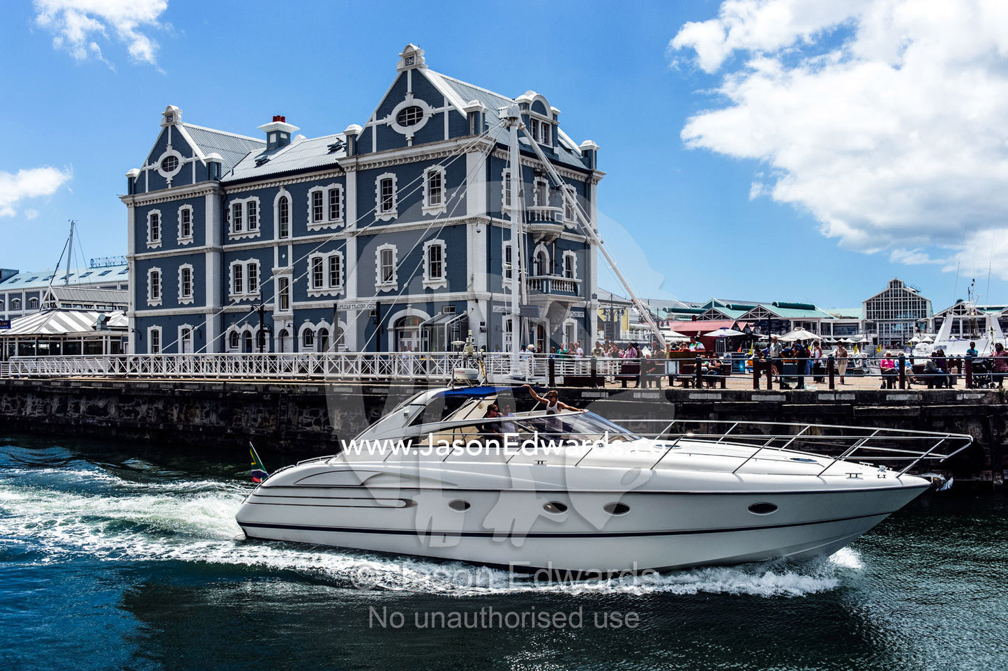 A luxury boat sails through a marina and docks crowded with tourists. Cape Town, Victoria and Alfred Waterfront, Western Cape, South Africa.