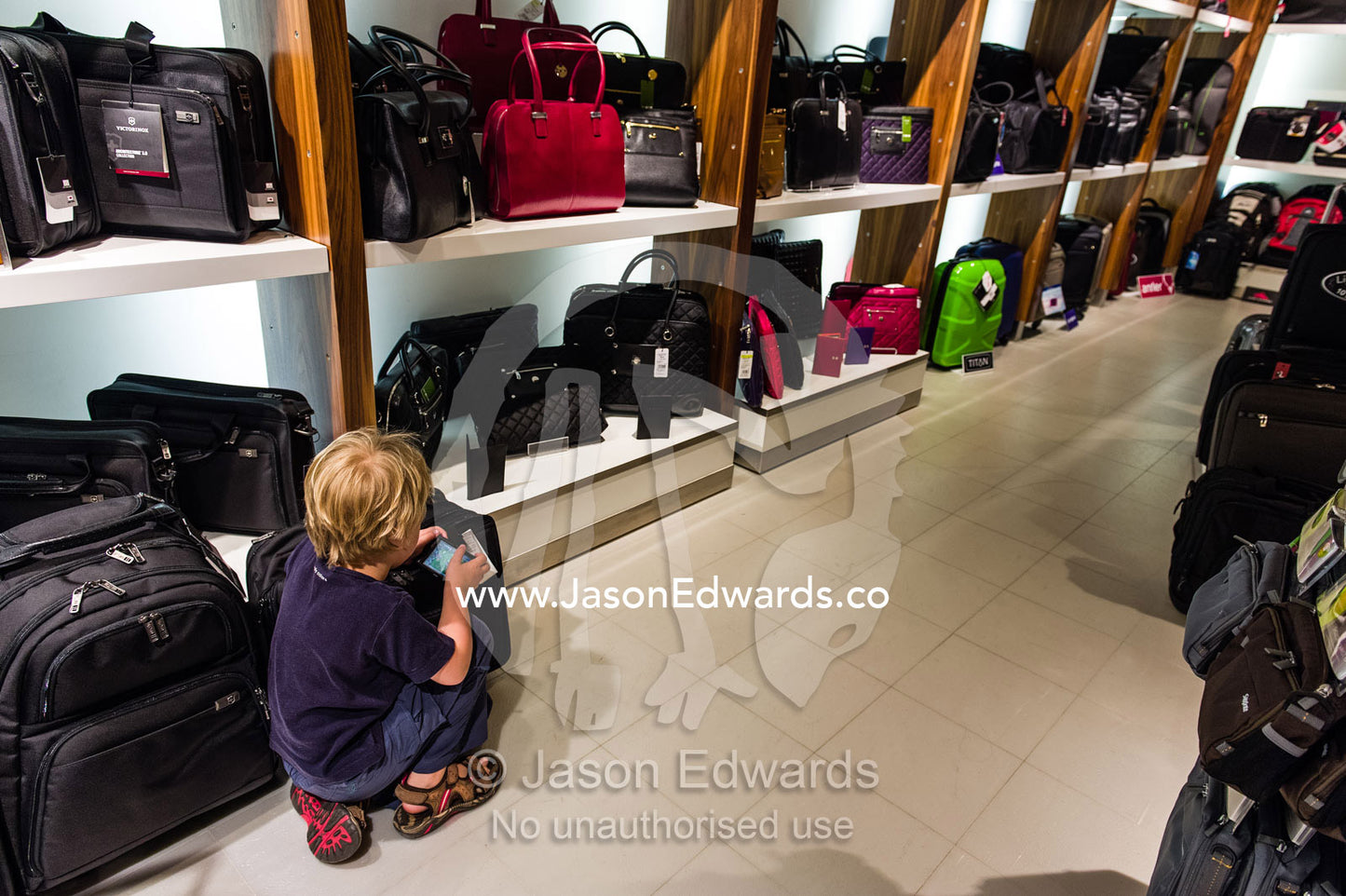A boy plays games on a cell phone whilst his parents buy a bag in an airport shop. Melbourne, Melbourne International Airport, Tullamarine, Victoria, Australia.