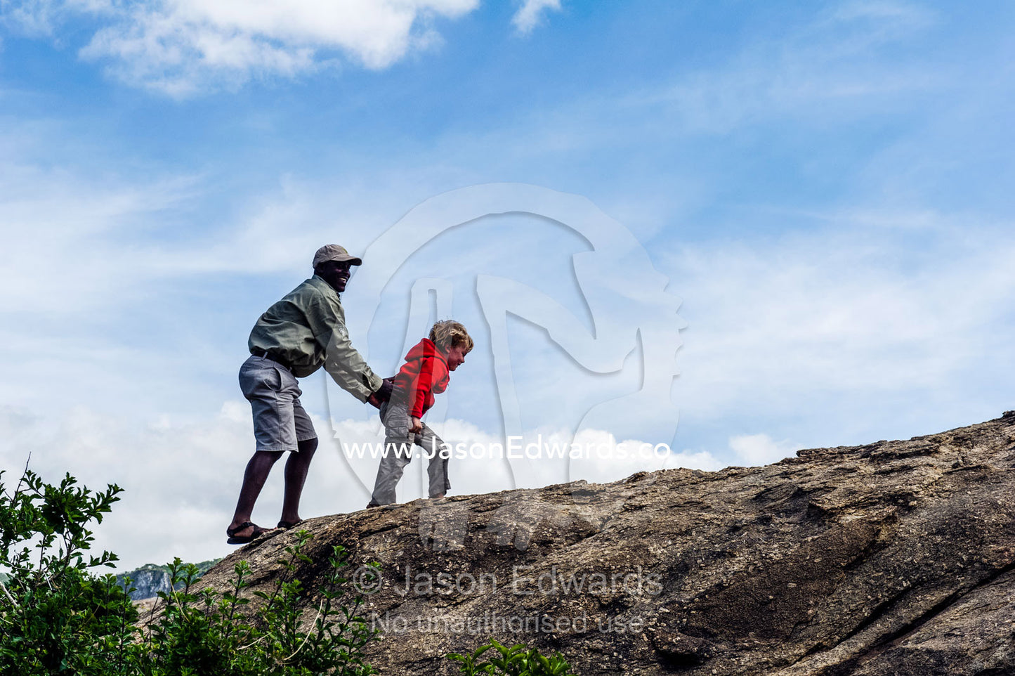 A guide helps a young boy on safari climb a granite outcrop known as a kopje on the savannah. Ngorongoro Conservation Area, Ngorongoro District, Tanzania, Africa.