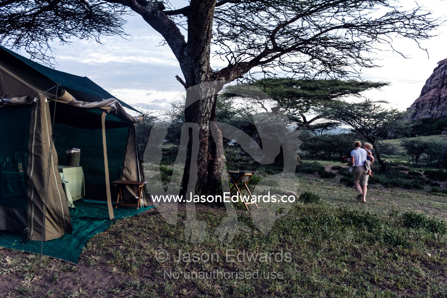 A mother and son walking through their campsite and dining tent whilst on safari. Arusha, Nasera Rock, Angata Kiti Valley, Ngorongoro Conservation Area, Crater Highlands, Tanzania, Africa.