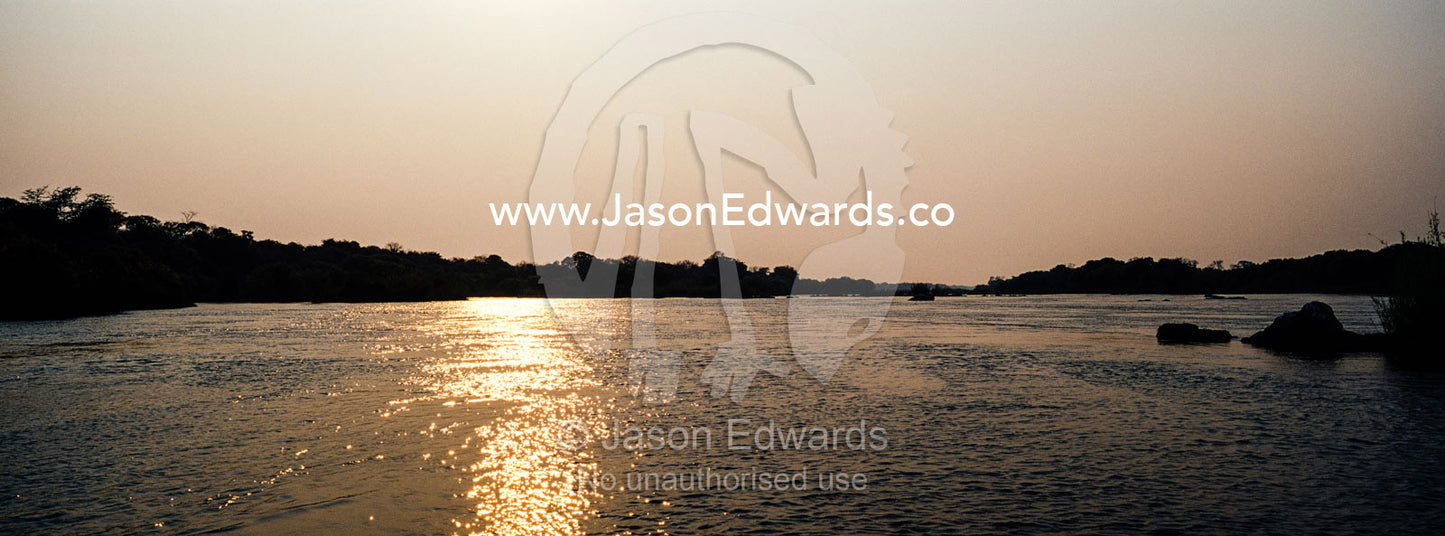 Dawn rises over a wide river flowing between riverbanks covered in forest. Chobe River, Chobe National Park, Botswana.