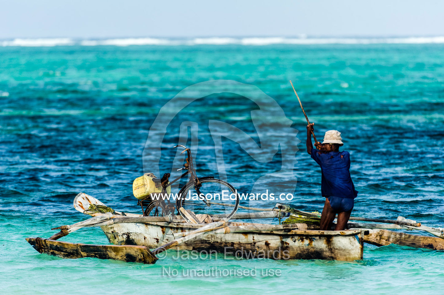 A fisherman poles his wooden trimaran dhow and bicycle through the shallows towards a reef. Bwejuu, Echo Beach Hotel, Echo Beach, Unguja Island, Zanzibar, Tanzania, Africa.
