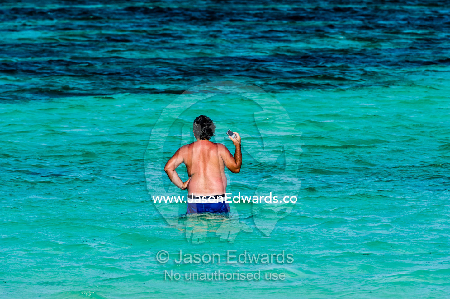 A tourist wading in the shallow, turquoise waters of the Indian Ocean with his pocket camera. Bwejuu, Echo Beach Hotel, Echo Beach, Unguja Island, Zanzibar, Tanzania, Africa.