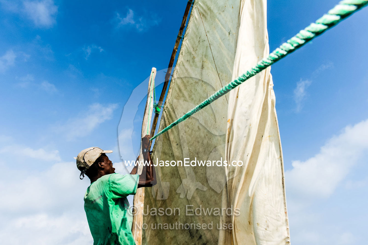 A deckhand on a wooden trimaran dhow working a tarpaulin sail under a blistering sun. Bwejuu, Unguja Island, Zanzibar, Tanzania, Africa.