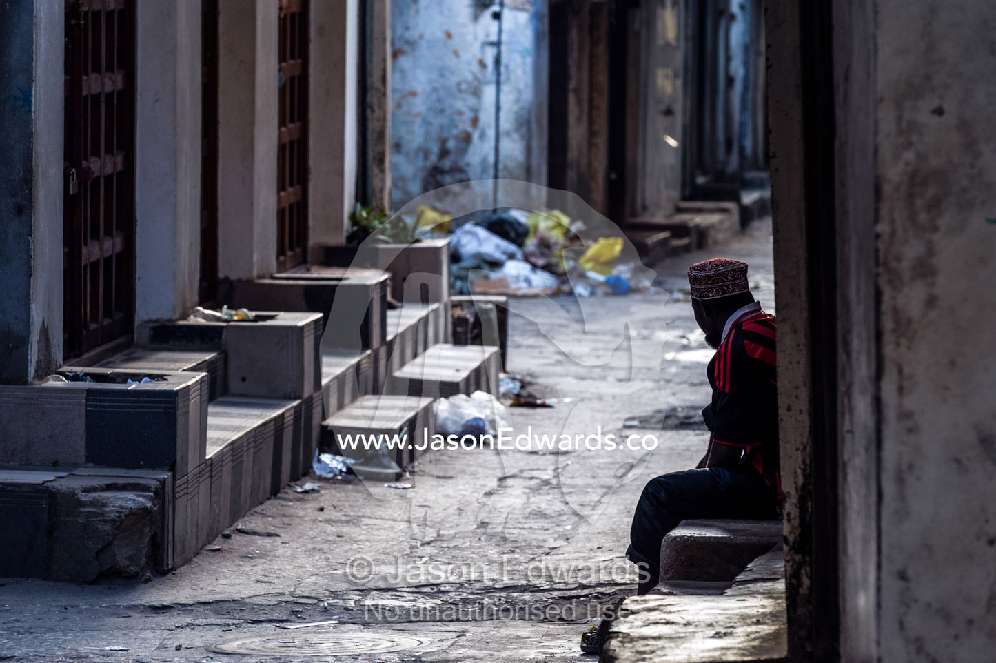 At dawn a Muslim man rests on a step in a narrow alley waiting for shops to begin trading. Stone Town, Mji Mkongwe, Zanzibar, Unguja Island, Zanzibar, Tanzania.