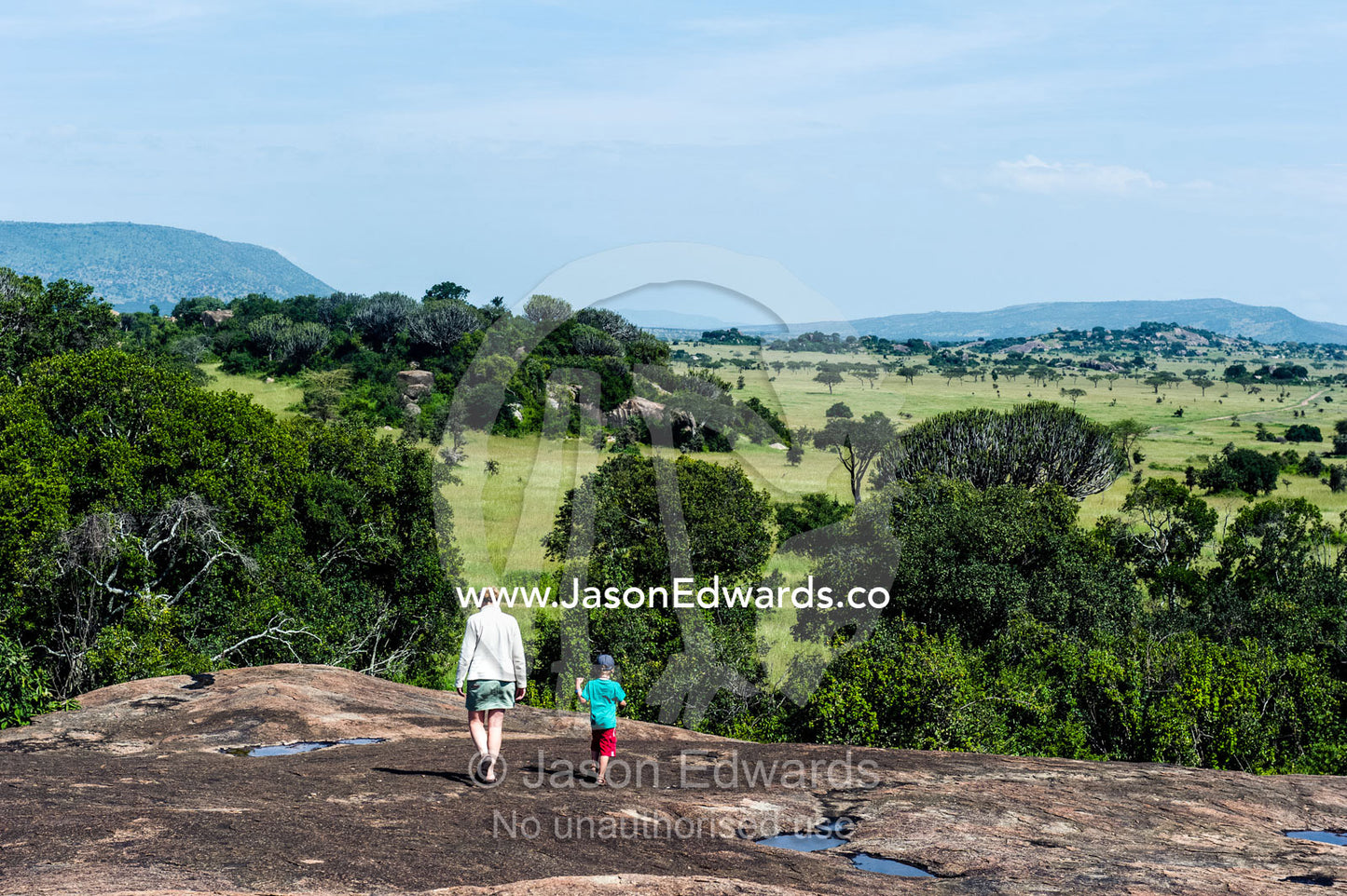 A mother and son hiking across the top of a granite kopje above a savannah plain. Moru Kopjes, Serengeti National Park, Serengeti, World Heritage Site, Tanzania.