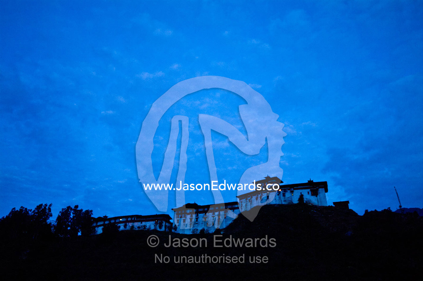 Lamp light peeks from windows atop the high walls of an ancient Buddhist monastery on a mountaintop at night. Punakha River, Punakha Valley, Punakha-Wangdiphodrang District, Bhutan.