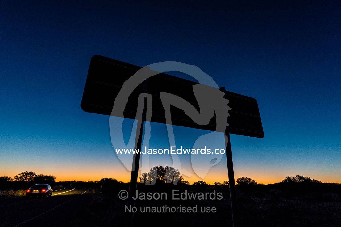A car passes a directional sign silhouetted on a remote outback desert road at sunset. Uluru Kata Tjuta National Park, Northern Territory, Australia.