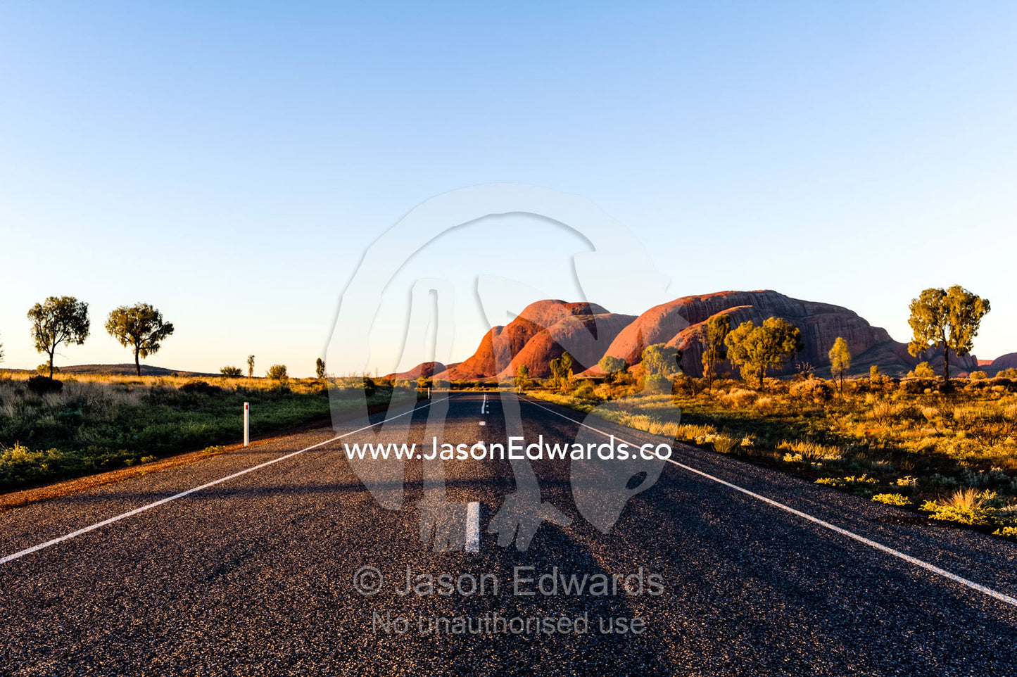 A bitumen road crosses the outback desert plain towards the domes of Kata Tjuta at sunset. Uluru Kata Tjuta National Park, Northern Territory, Australia.