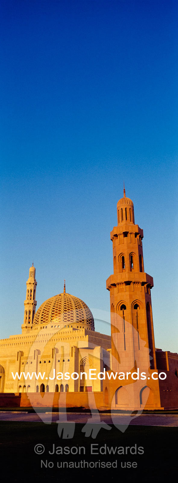 An ornate stone minarette towers over the dome of the Prayer Hall of the Grand Mosque at sunset. Sultan Qaboos Grand Mosque, Muscat, Sultanate of Oman.