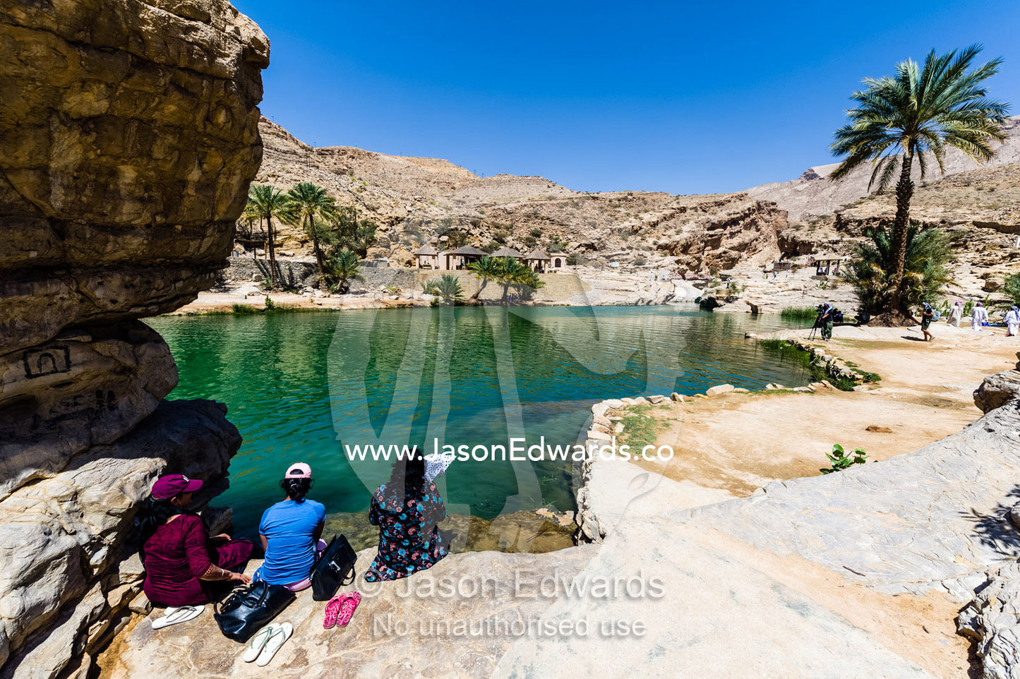 Tourists sit by the shore of the turquoise waters of a wadi in a desert valley. Wadi Bani Khalid, Sharqiyah Region, Sultanate of Oman.