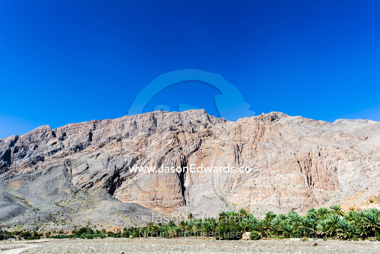 Palm trees follow the course of a river as it winds it's way along the base of a desert mountain. Adh Dhahir, Dima Wal Taeen, North Al Sharqiyah Governorate, Sultanate of Oman.