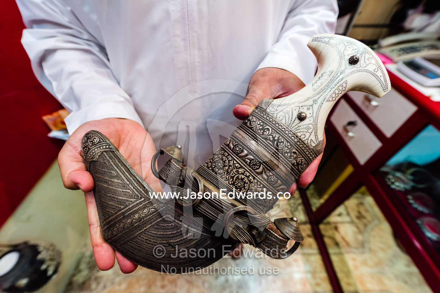 A salesman displays an expensive silver khanjar knif with an ivory handle for sale in a souq. Mutrah Souq, Muscat, Sultanate of Oman.