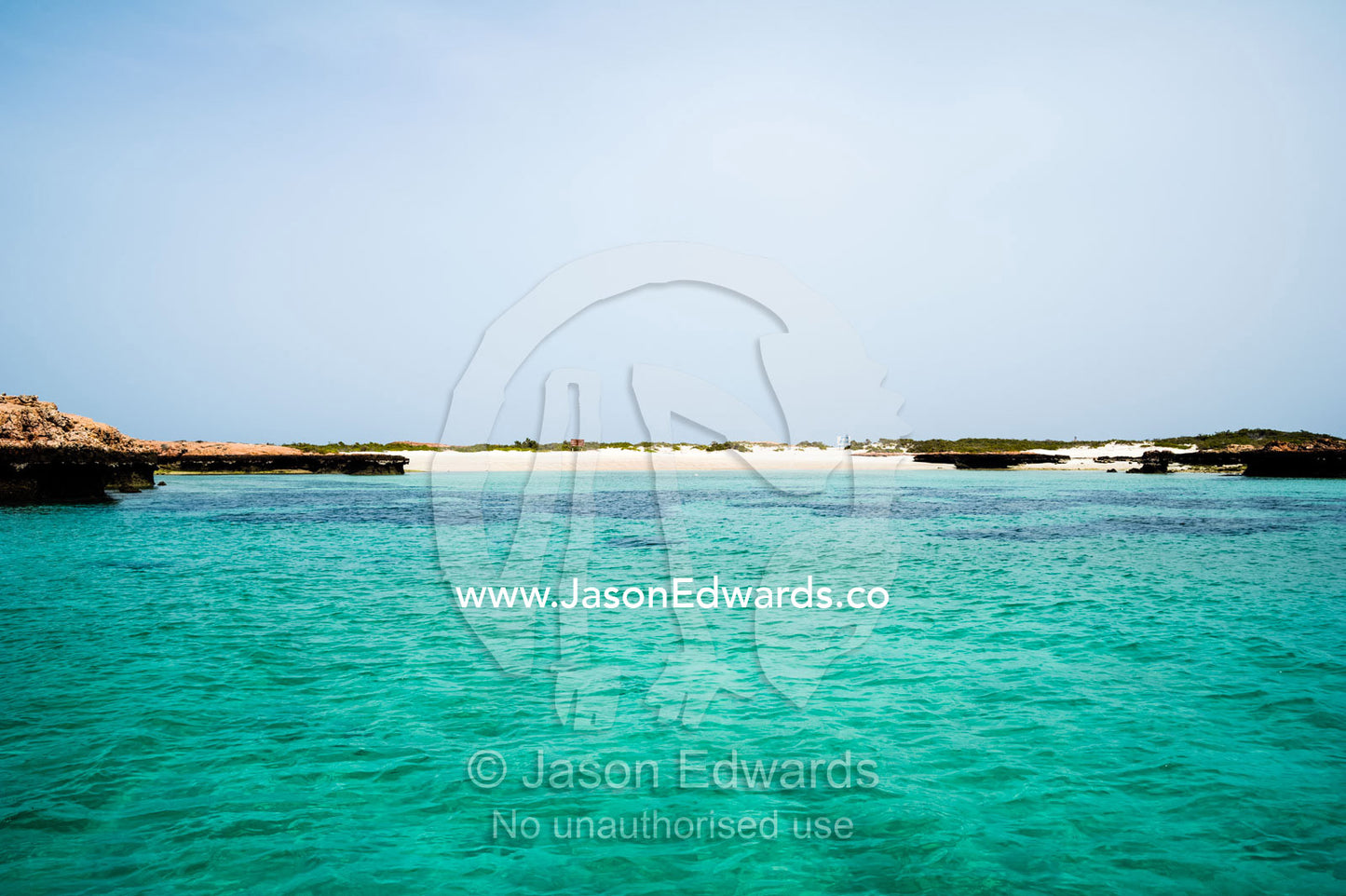 Clear, turquoise seas surround a barren red sandy desert island. Daymaniyat Islands Nature Reserve, Wilayat AlSeeb, Barka, Sultanate of Oman.