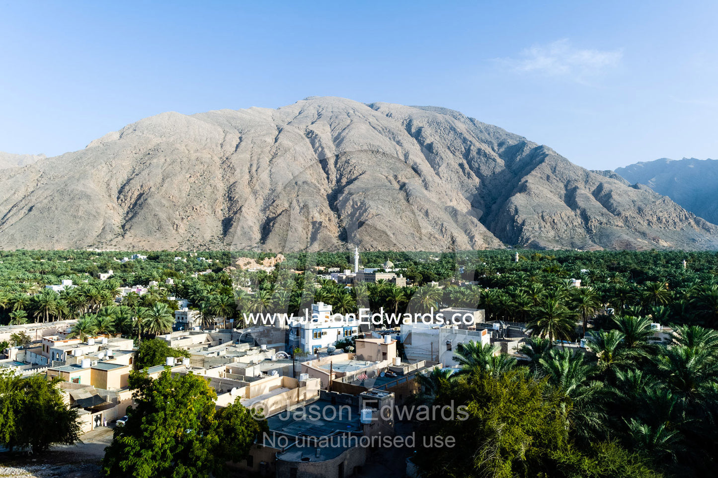 A date palm tree grove and village fills a desert valley floor beneath jagged mountain peaks. Wilayt Nakhal, Mount Nakhal, Al Batinah South Governorate, Sultanate of Oman.