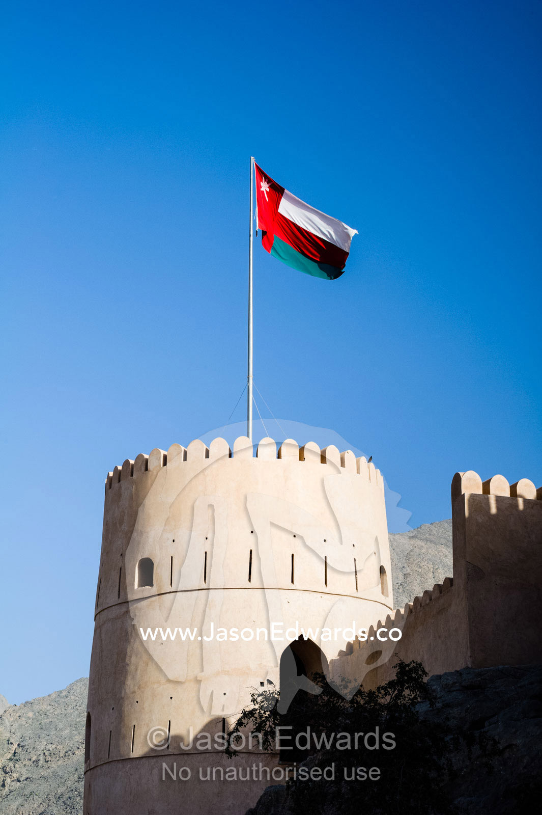 The Oman flag flies over the parapets and watch tower of a fort on a desert mountaintop. Nakhal Fort, Wilayt Nakhal, Mount Nakhal, Al Batinah South Governorate, Sultanate of Oman.