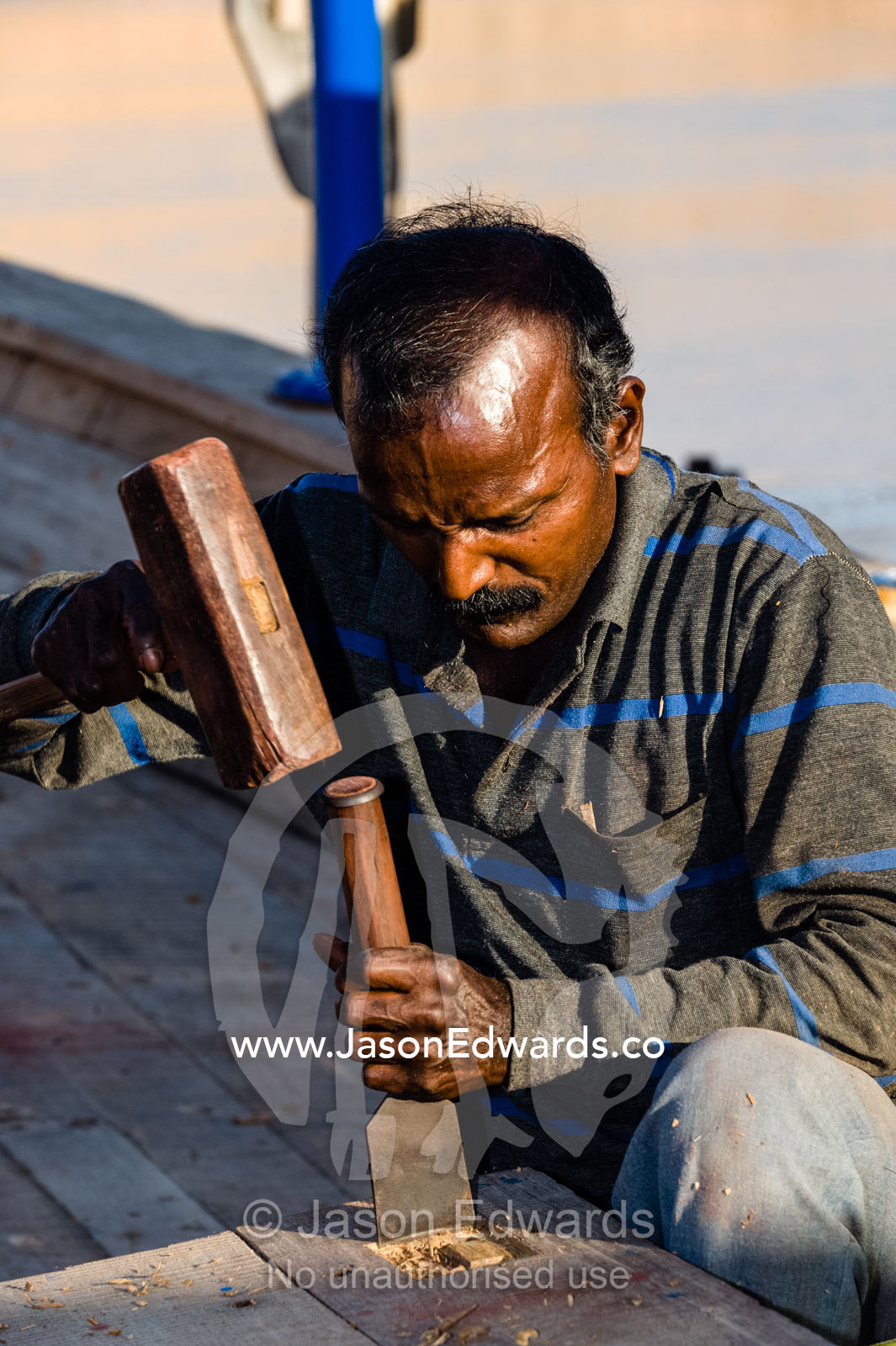 A laborer shapes timber with a hammer and chisel in a dhow ship building yard. Sur, Ash Sharqiyah Region, Gulf of Oman, Sultanate of Oman.